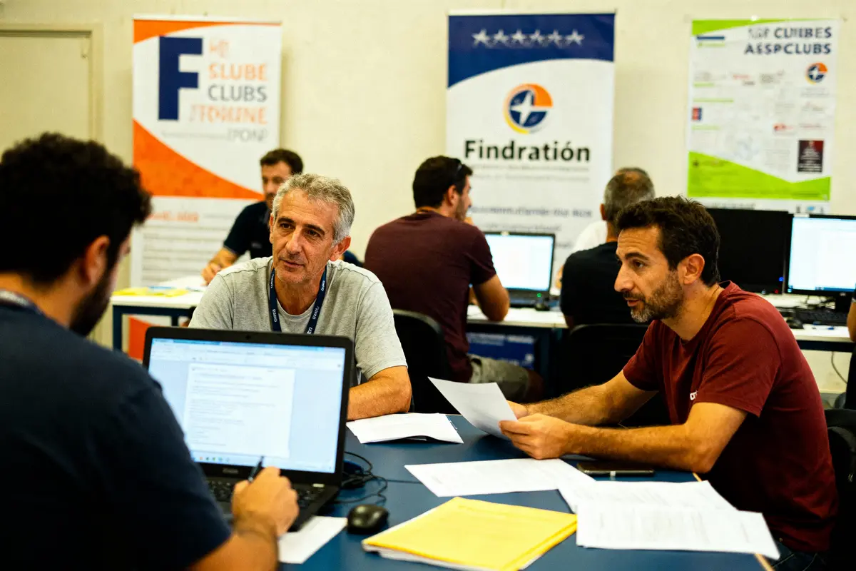 Des personnes discutent autour d'une table avec des ordinateurs dans un cadre professionnel, affiches au fond.