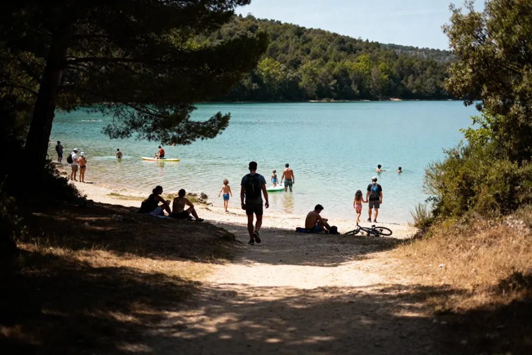 Gens profitant de la plage, forêt autour, eau turquoise, ambiance détendue, ombre, vélo posé au sol.