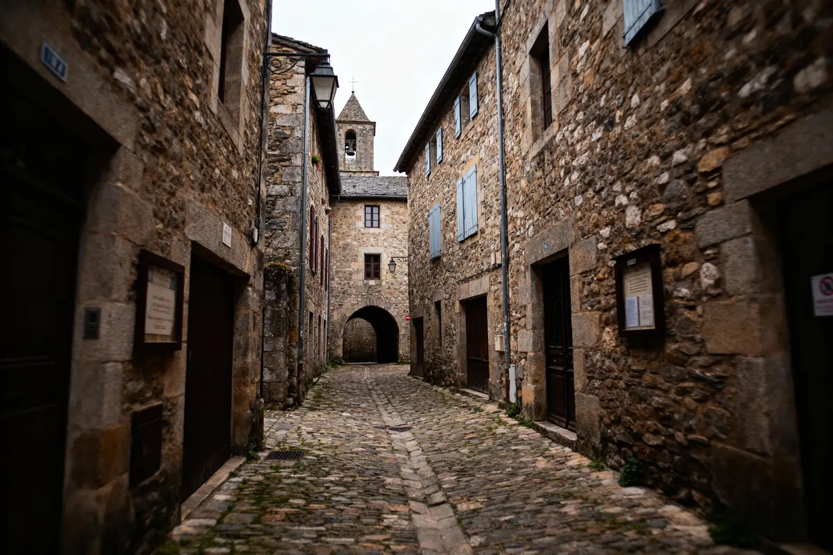 Rue pavée étroite, bâtiments en pierre, volets bleus, arche, clocher. Ambiance médiévale, ciel nuageux.