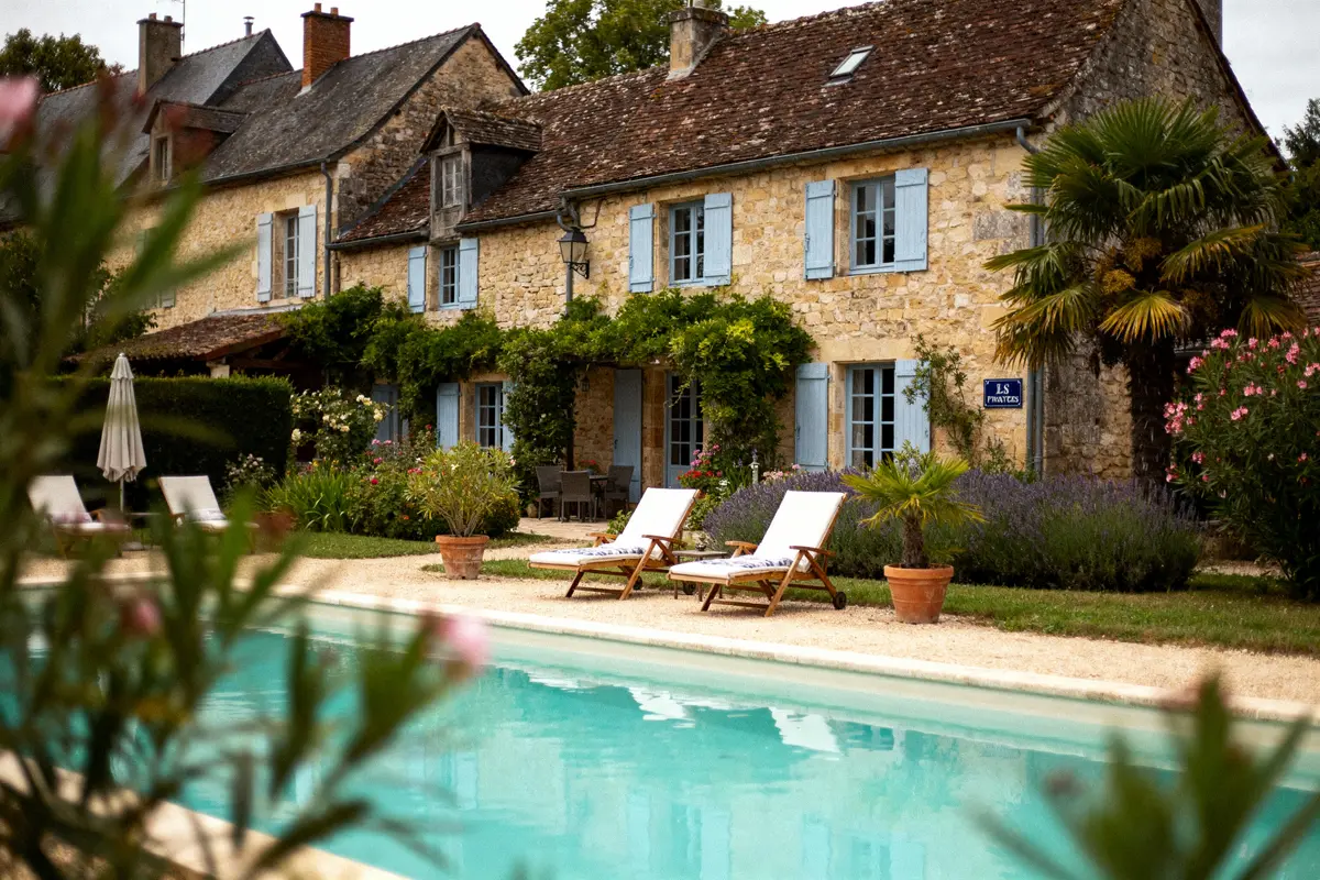 Maison en pierre avec volets bleus, piscine, chaises longues, végétation, cadre estival et ciel couvert.