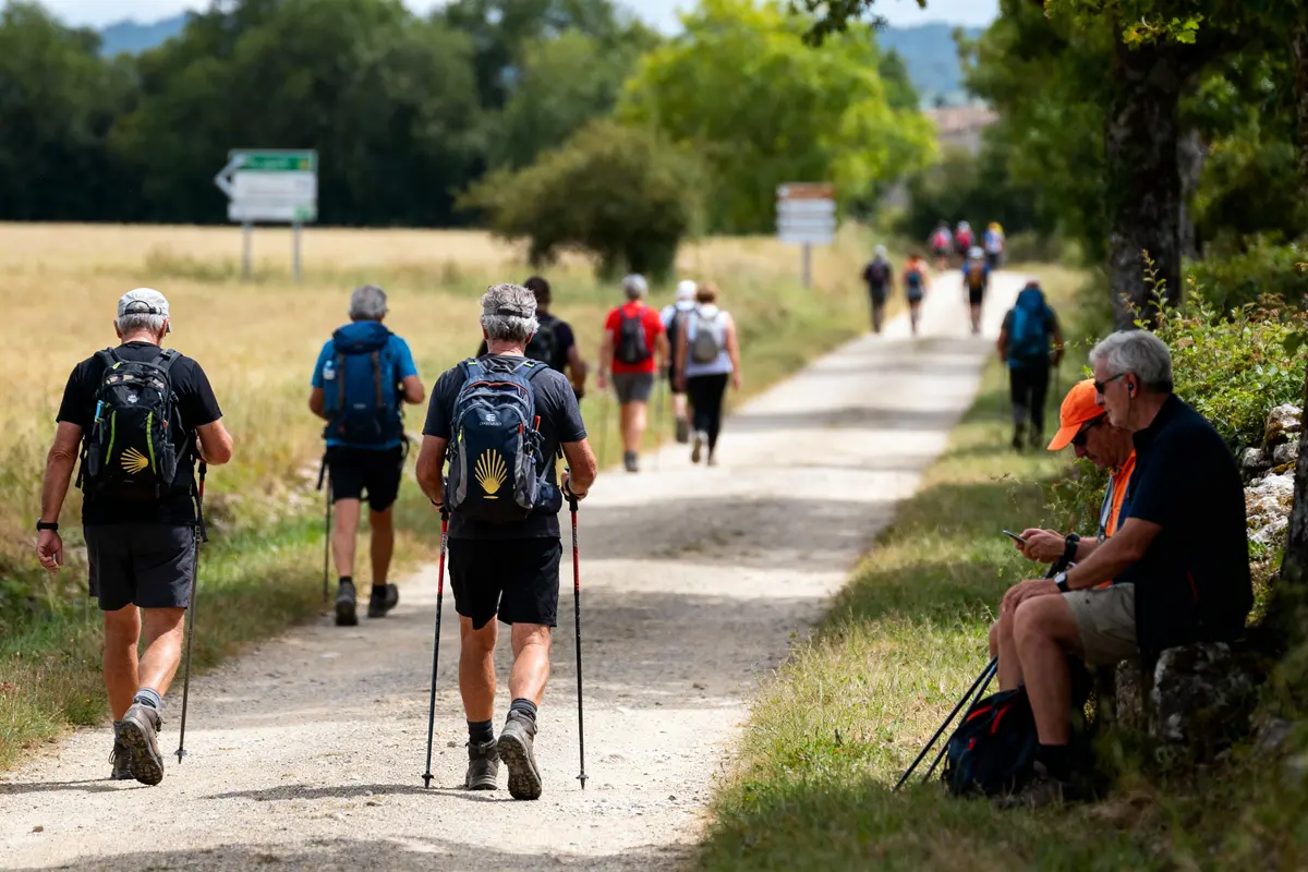 Groupe de randonneurs marchant sur un chemin, deux hommes assis sur le côté, paysage verdoyant.