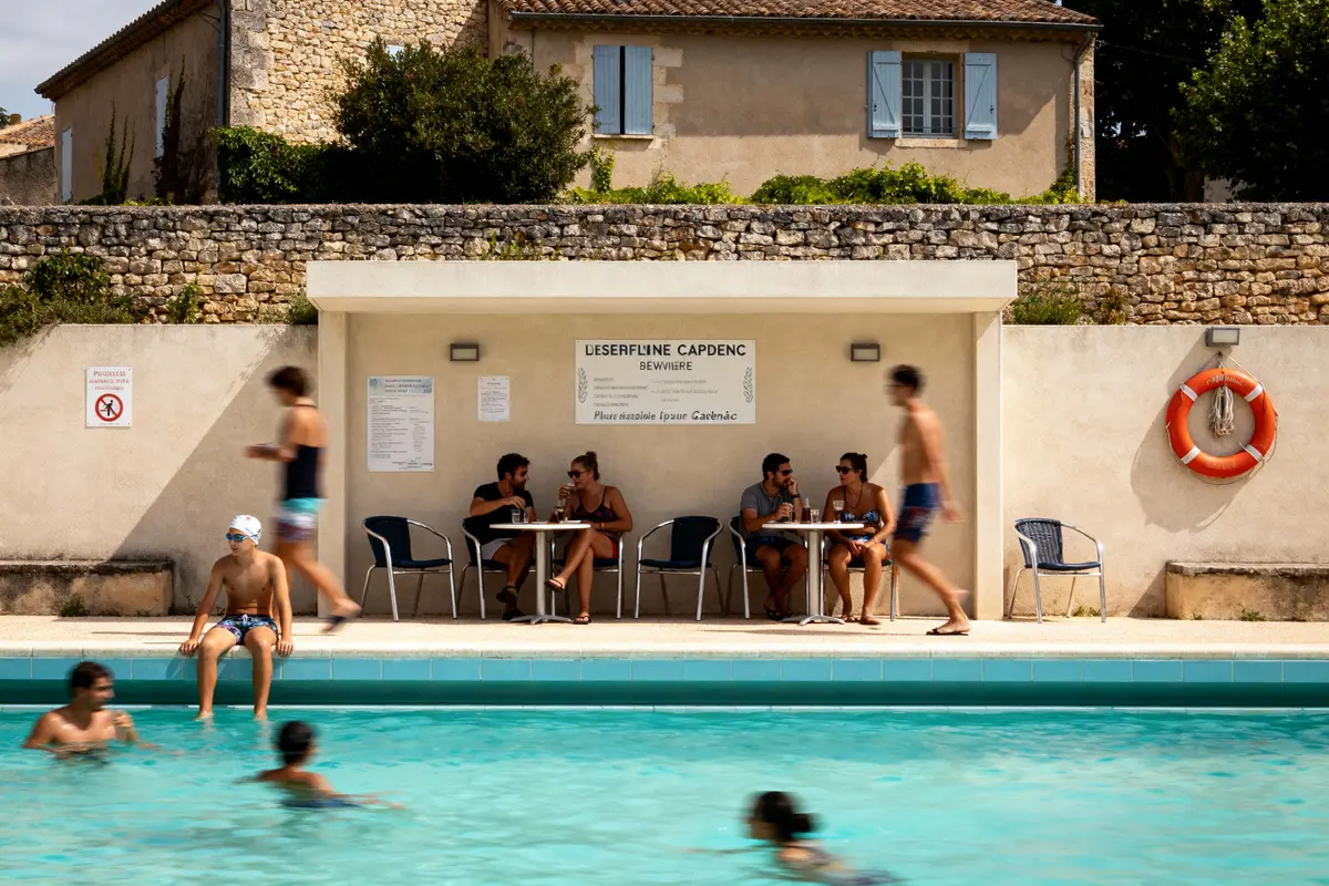Piscine extérieure avec personnes assises à une table et nageurs, bâtiment en pierre et bouée en arrière-plan.