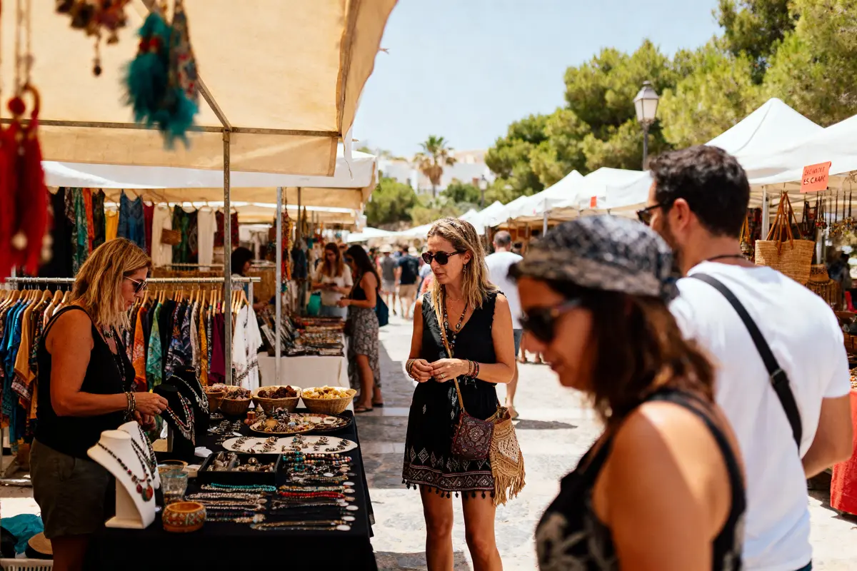 Marché en plein air, femmes examinant des bijoux artisanaux sous des tentes blanches, journée ensoleillée, ambiance vivante.