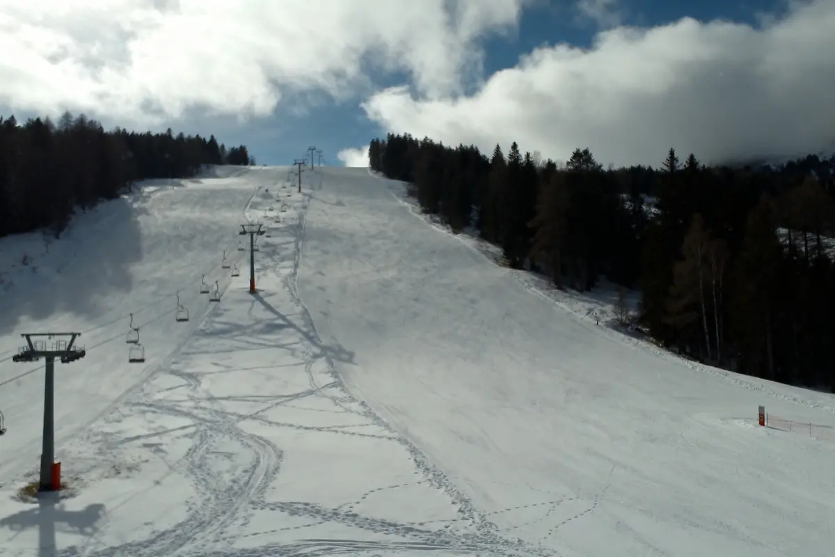 Piste de ski enneigée avec télésiège, entourée de sapins, sous ciel nuageux et bleu.