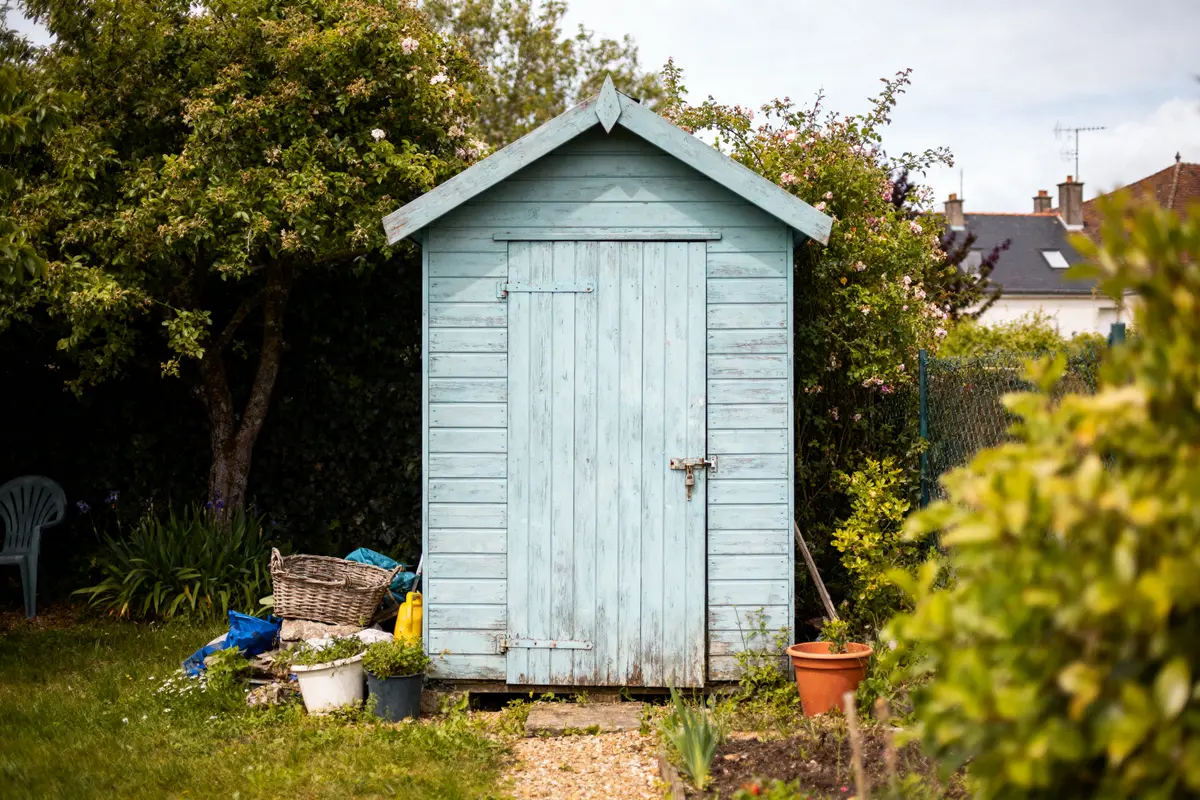 Abri de jardin en bois bleu, entouré de plantes vertes, pot et panier devant.