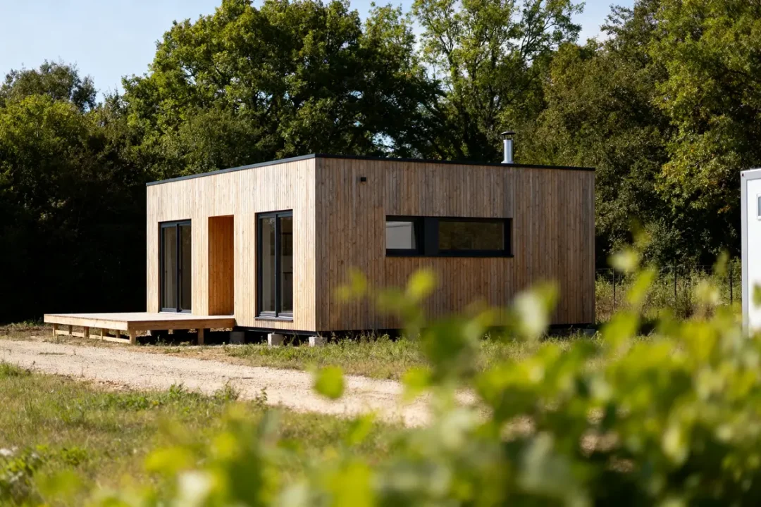 Petite maison en bois moderne avec terrasse, entourée de verdure et d'arbres, par une journée ensoleillée.