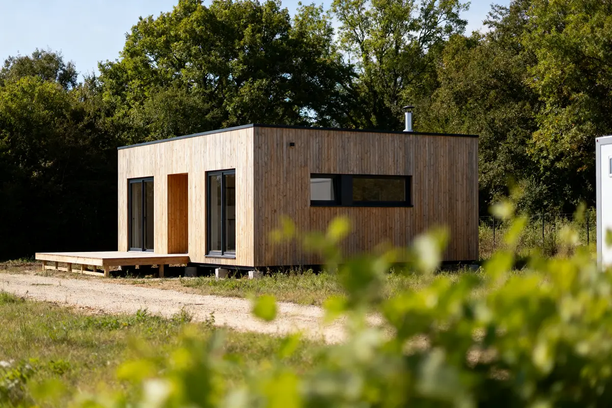 Petite maison en bois moderne avec terrasse, entourée de verdure et d'arbres, par une journée ensoleillée.