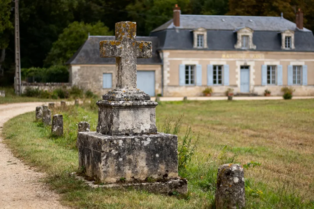 Croix en pierre ancienne au premier plan, maison en arrière-plan, chemin et pelouse verdoyante.