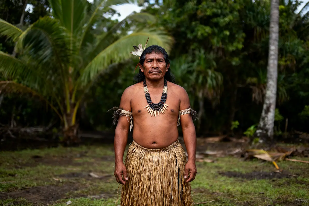 Homme indigène en tenue traditionnelle, collier et coiffe, debout dans une jungle luxuriante.
