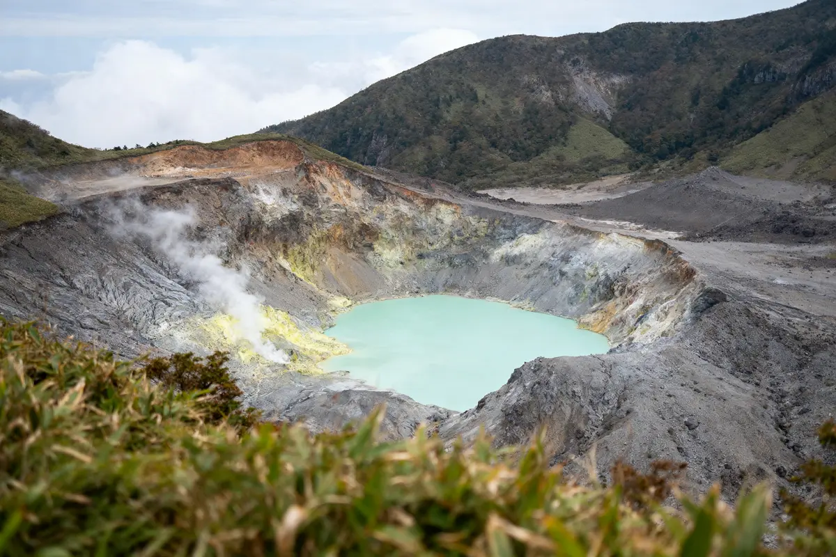 Cratère volcanique avec lac turquoise, montagnes environnantes, buissons au premier plan, vapeur s'élevant du sol.