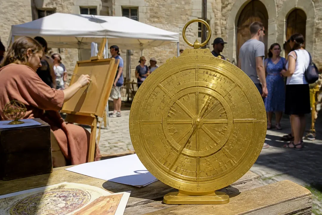 Image d'un astrolabe doré sur table, avec une femme dessinant en arrière-plan, lors d'un événement extérieur.