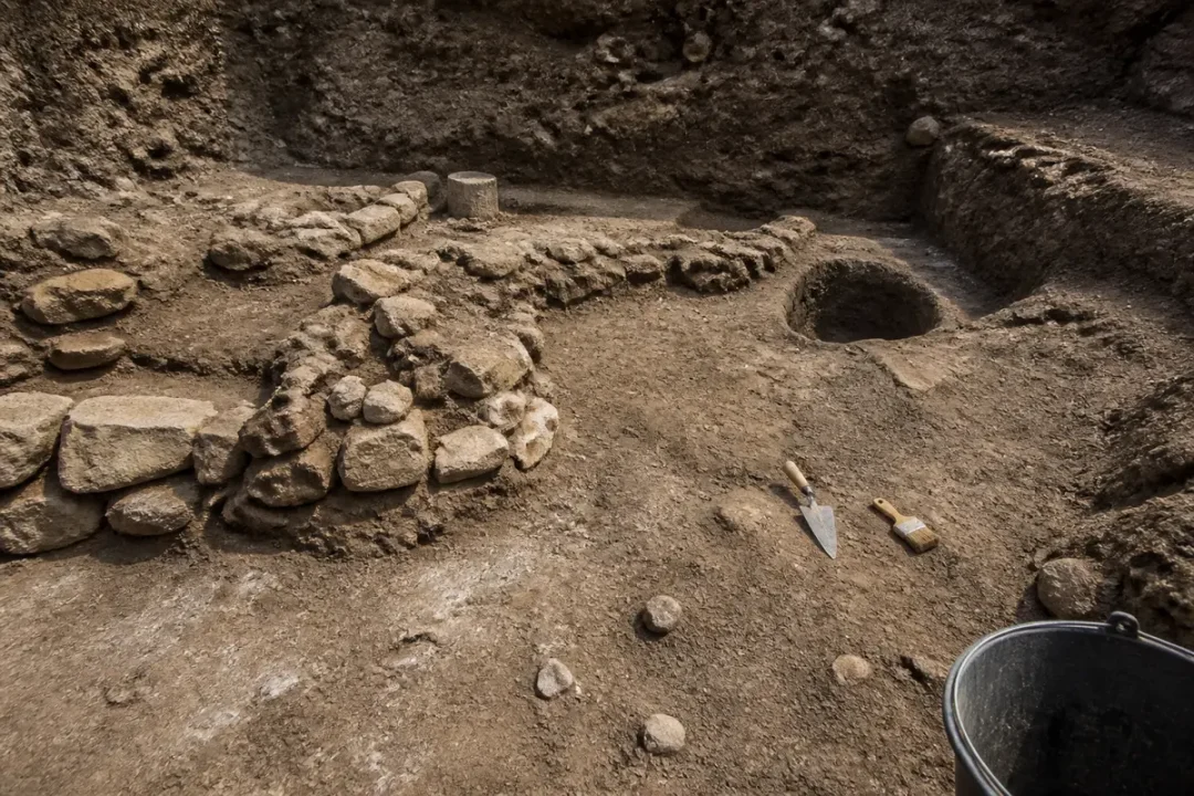 Site de fouilles archéologiques avec pierres, outils et seau sur un sol terreux.
