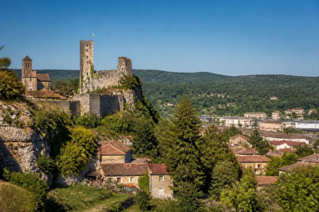 Ancien château médiéval en ruines surplombant village pittoresque, arbres verdoyants, colline et ciel dégagé.