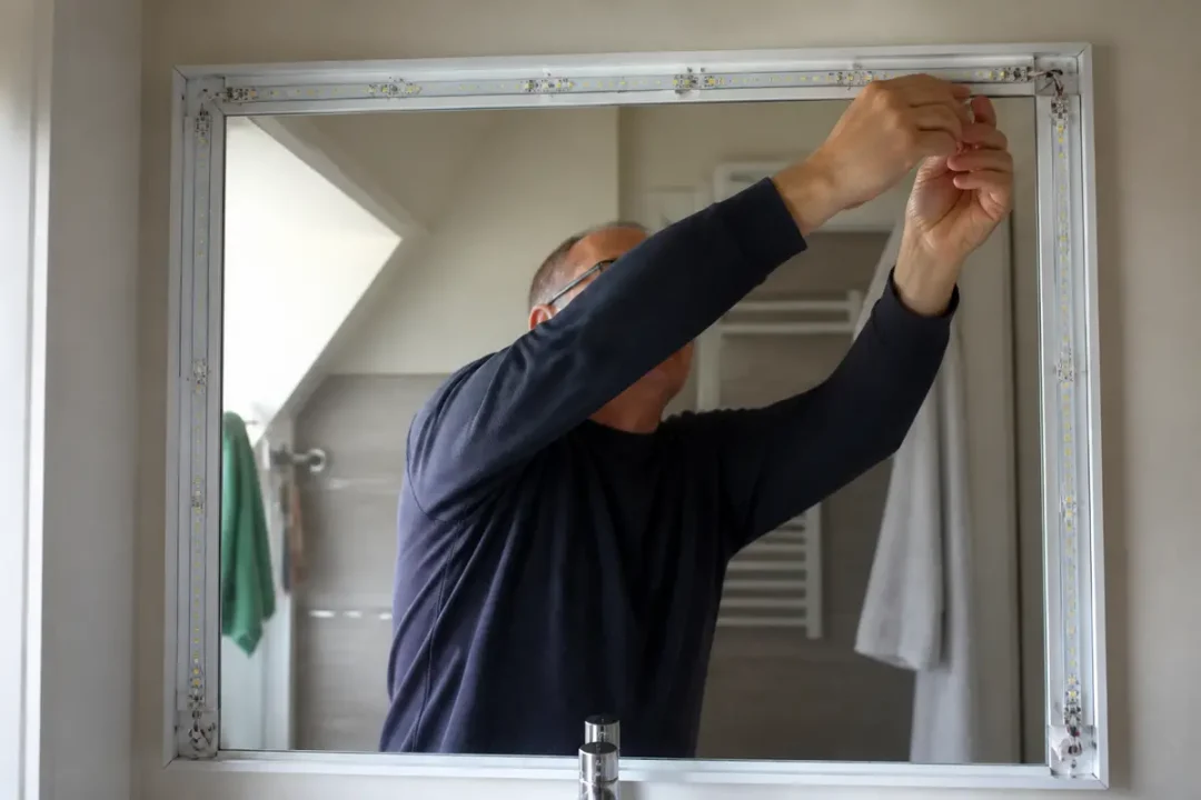 Homme installant un miroir lumineux dans une salle de bain moderne avec des serviettes accrochées.