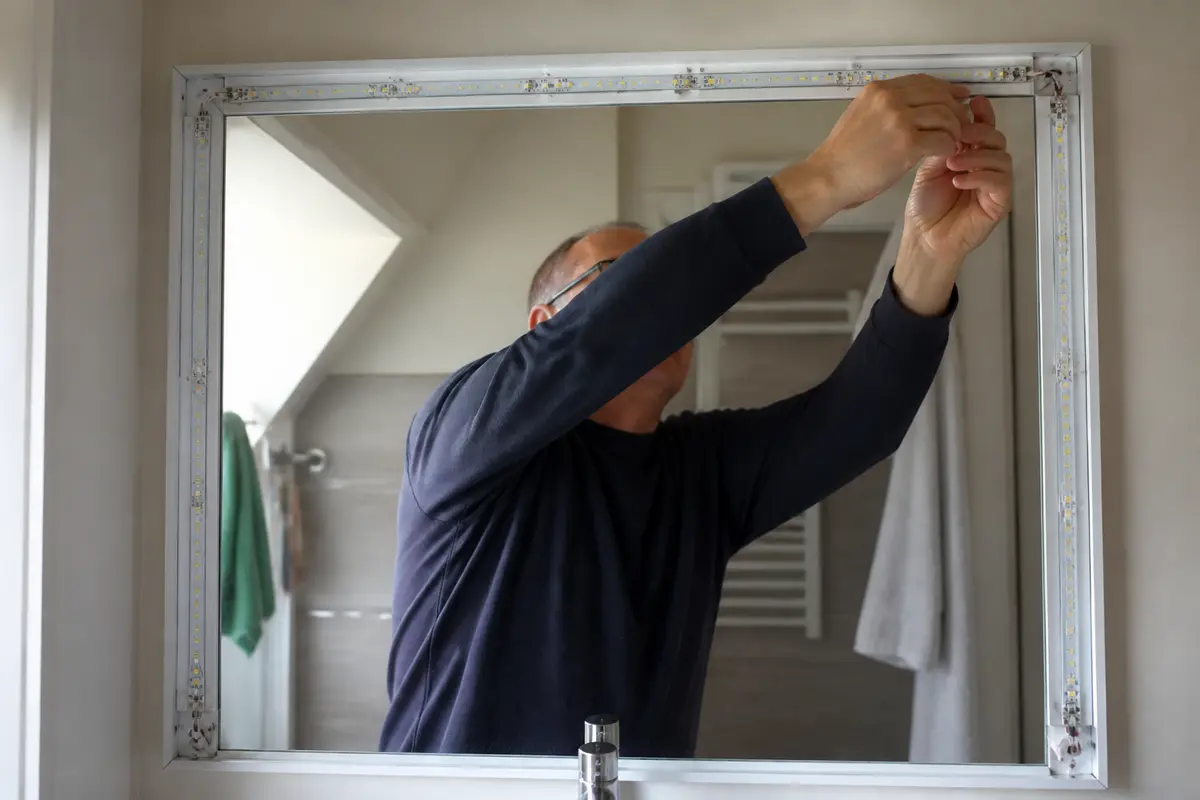 Homme installant un miroir lumineux dans une salle de bain moderne avec des serviettes accrochées.