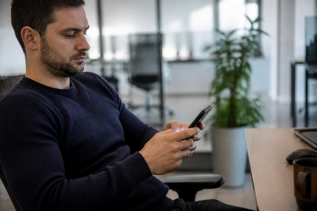 Homme assis utilisant un smartphone dans un bureau moderne, avec plante et bureau en arrière-plan.