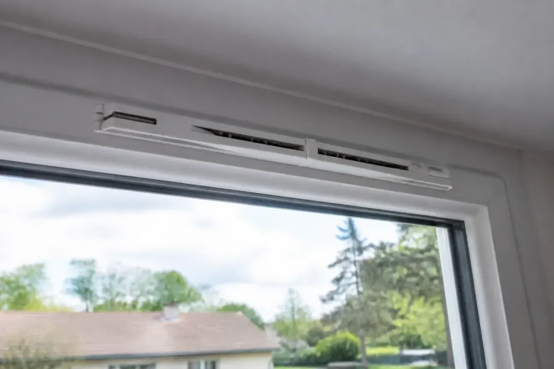 Grille d’aération blanche installée sur une fenêtre moderne, vue extérieure avec ciel et arbres verdoyants.