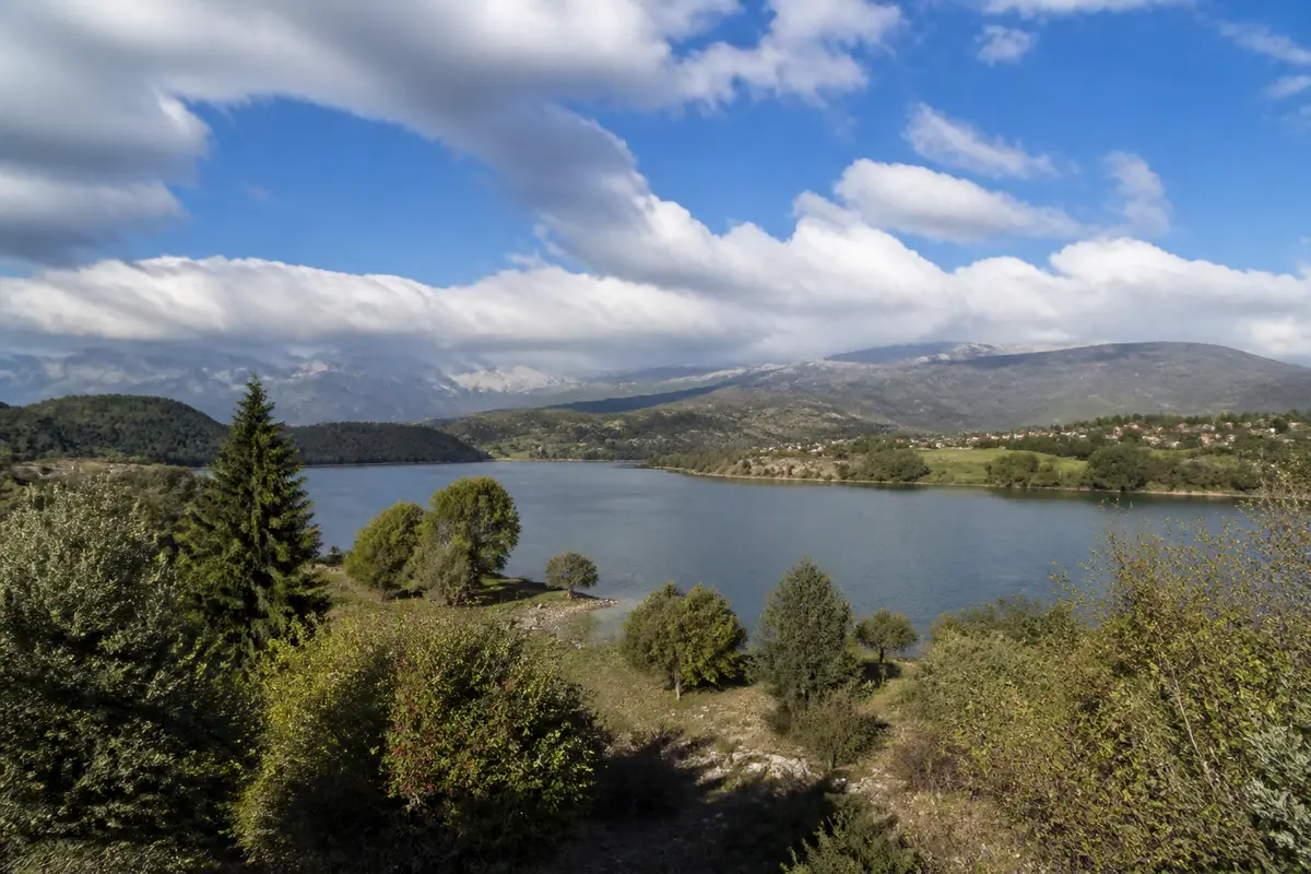 Lac entouré de végétation, collines verdoyantes et ciel partiellement nuageux. Paysage naturel calme et serein.
