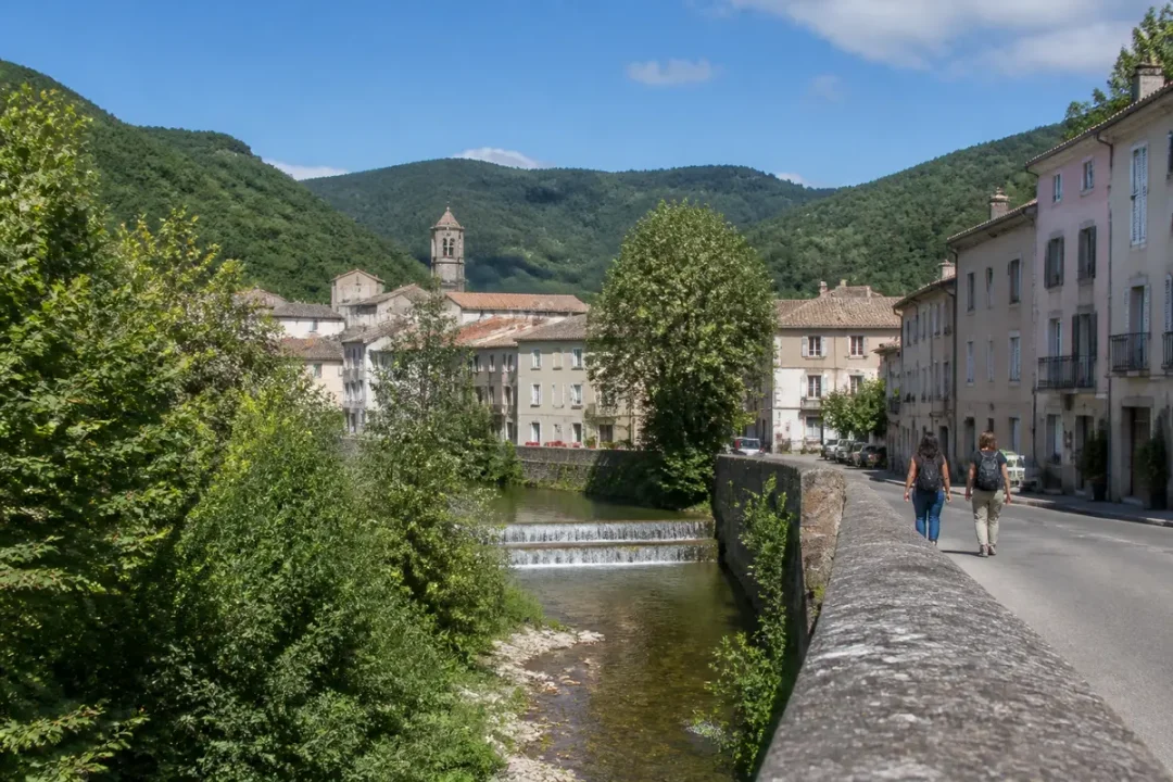Petite rivière traversant un village avec église, entourée de montagnes verdoyantes et ciel bleu.