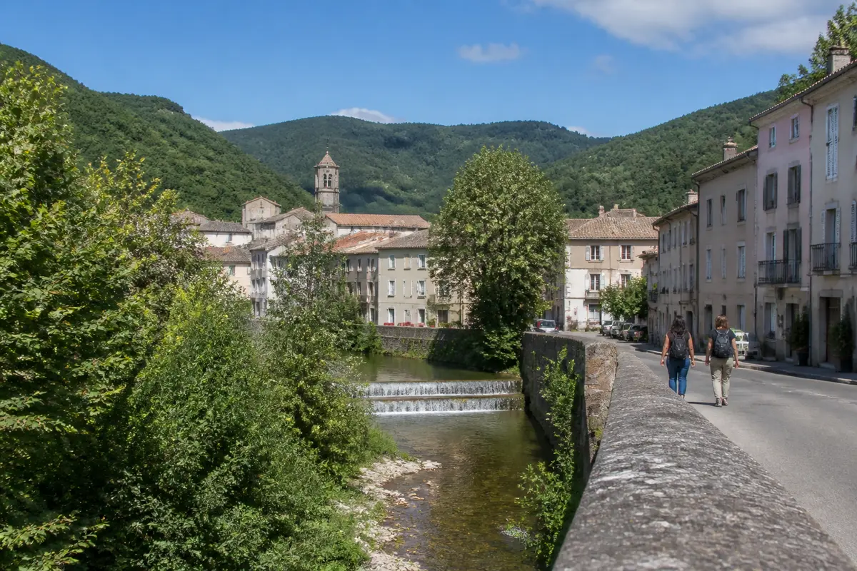 Petite rivière traversant un village avec église, entourée de montagnes verdoyantes et ciel bleu.