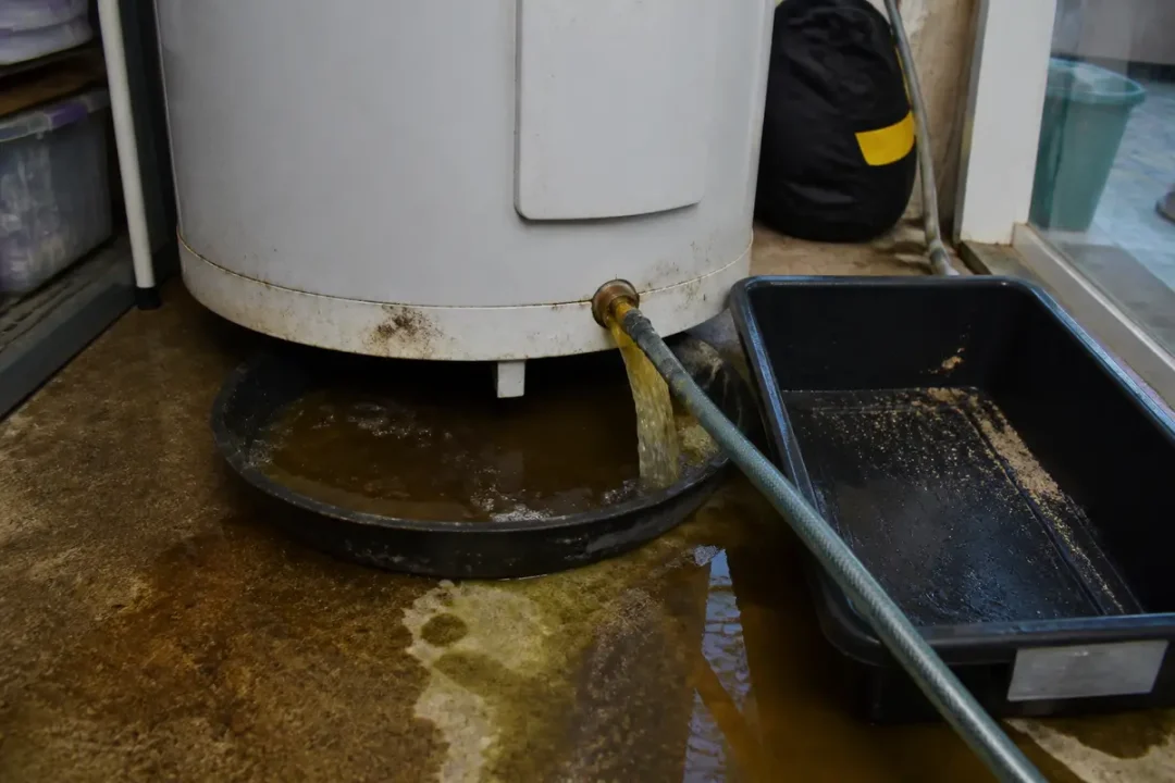 Chaudière purgeant de l'eau brune par un tuyau dans un seau, sol cimenté humide.